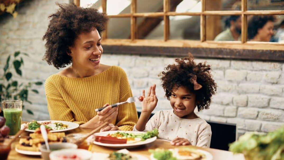 A woman and her young daughter sit the dinner table. The daughter is refusing a vegetable from her mom.