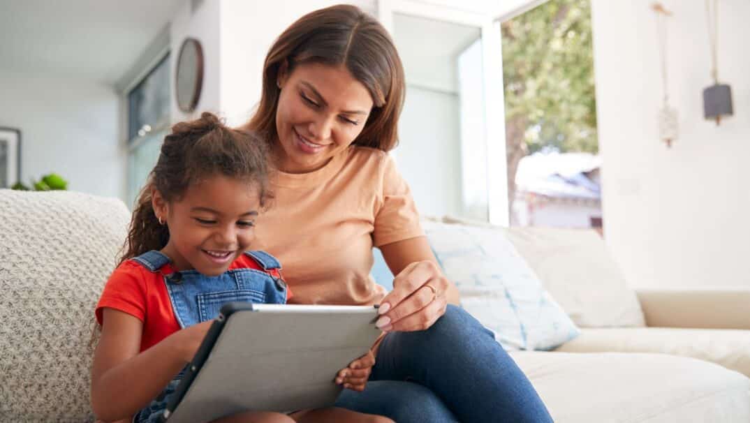 Mother and young daughter sit on the couch using a tablet together.