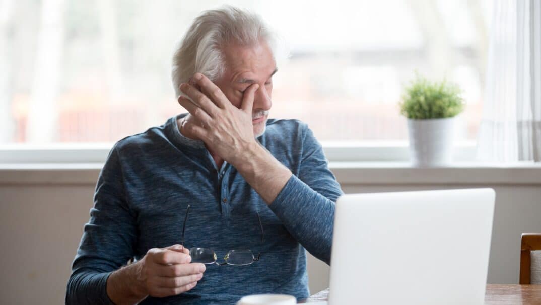 Older white man rubbing his eyes, holding his glasses, while sitting in front of his laptop.