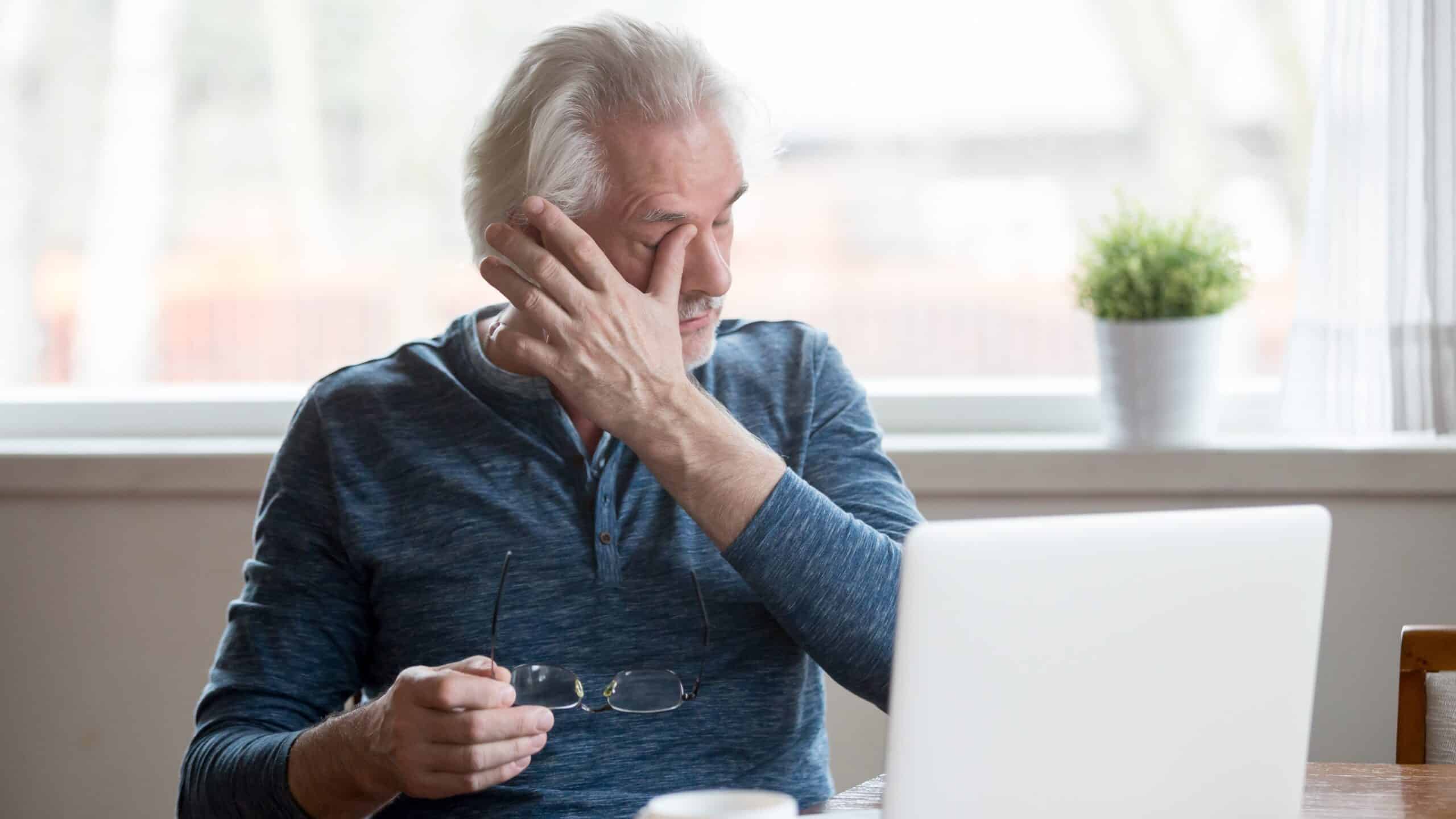 Older white man rubbing his eyes, holding his glasses, while sitting in front of his laptop.