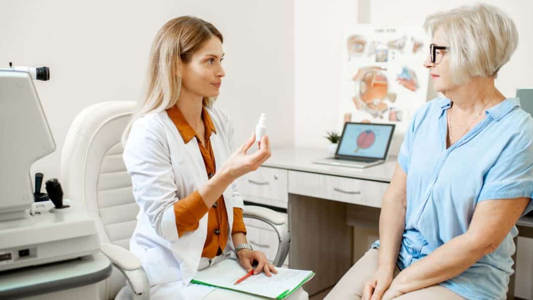 An ophthalmologist sits with a patient in her office. The patient is a middle-aged woman, wearing a blue blouse and glasses. The ophthalmologist is holding a bottle of eye drops.