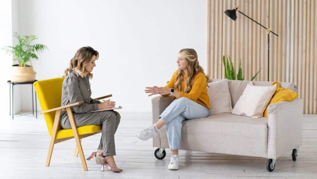 Young adult woman in an orange sweater sitting on a white sofa talking to a woman therapist with a clipboard. The therapist is wearing a gray suit.