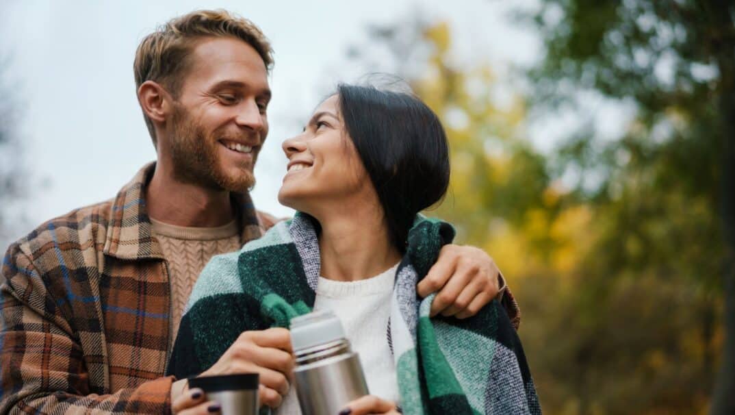 A young couple is outside, the man has his arms around the woman. She is holding a thermos.