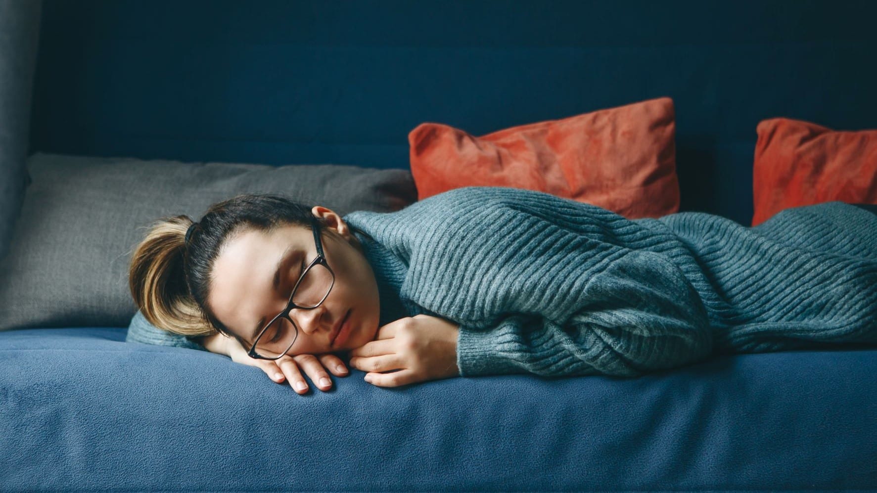 Young woman laying on a navy couch with her glasses still on. She is using her arm as a pillow, but there are orange pillows in the background of the image.