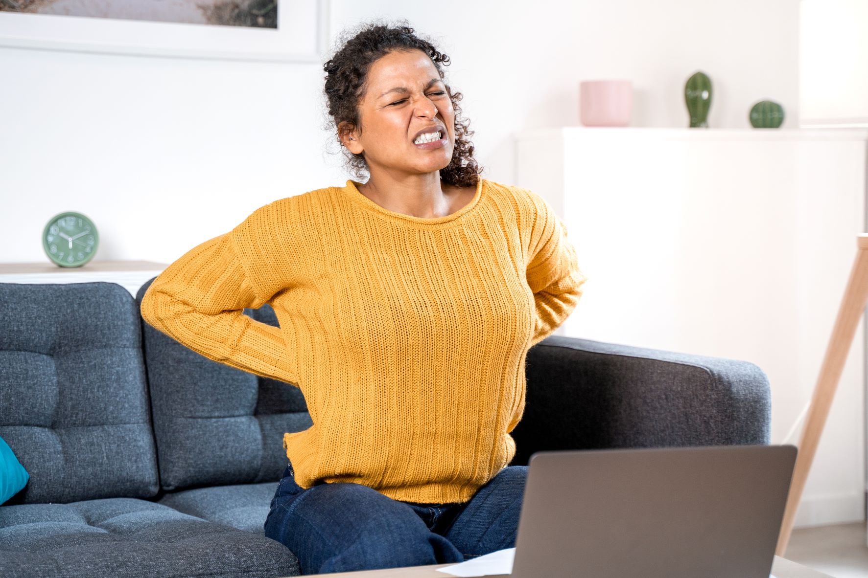 Woman sitting on a couch, holding her back and grimacing in pain.