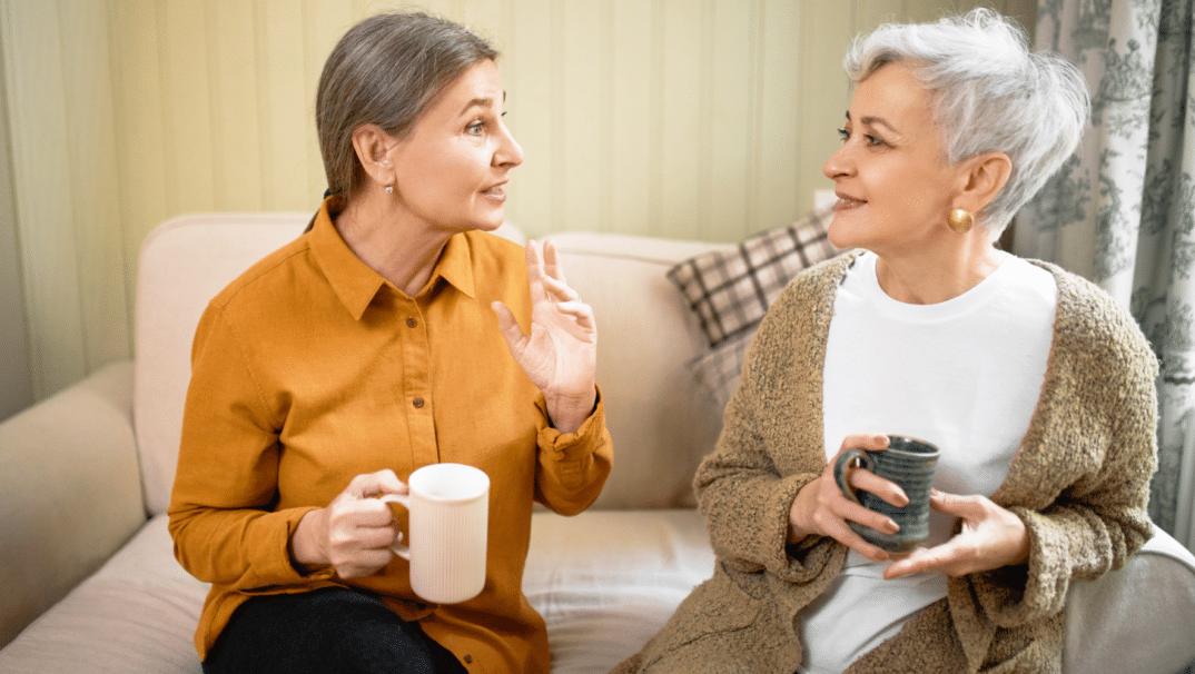 Woman with orange blouse sitting on sofa and drinking coffee with another woman with short hair wearing a green cardigan.